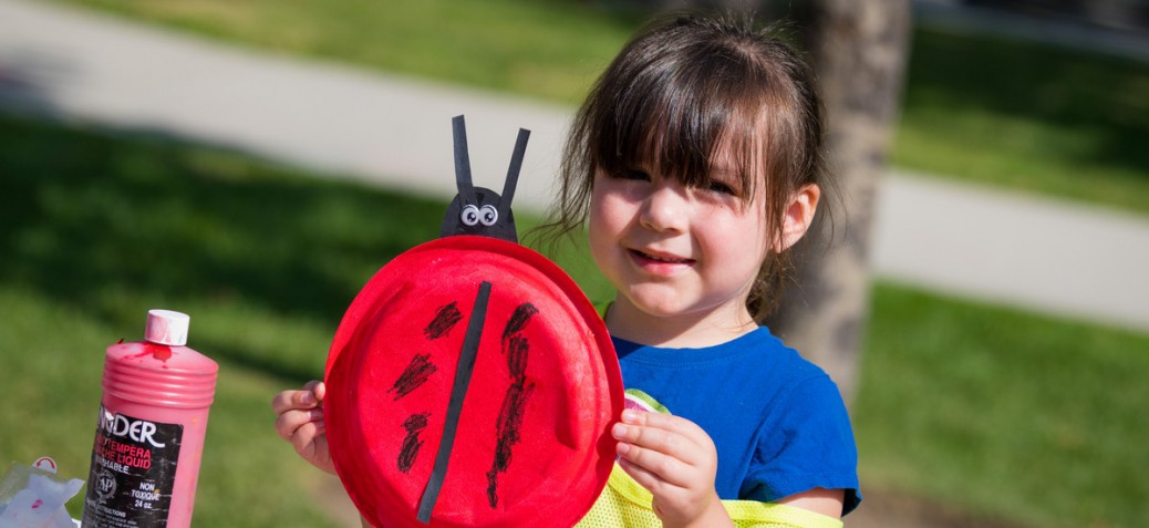 A young girl with dark hair and bangs holds up her ladybug art project and smiles at the camera.