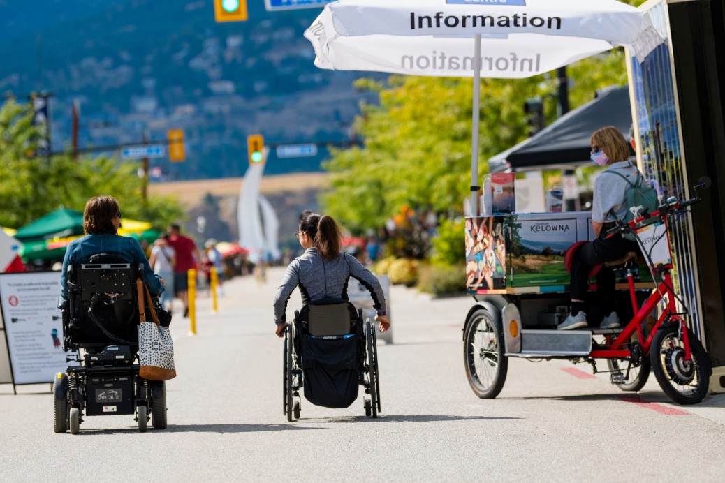 Two residents, one in a power chair and one in a wheelchair, rolling down Bernard Avenue during Meet me on Bernard on a sunny summer day.