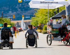 Two residents, one in a power chair and one in a wheelchair, rolling down Bernard Avenue during Meet me on Bernard on a sunny summer day.